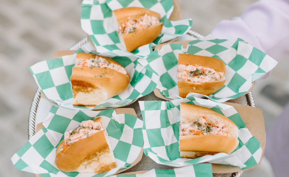 A tray holding six mini sandwiches on green and white checkered paper, served outdoors.