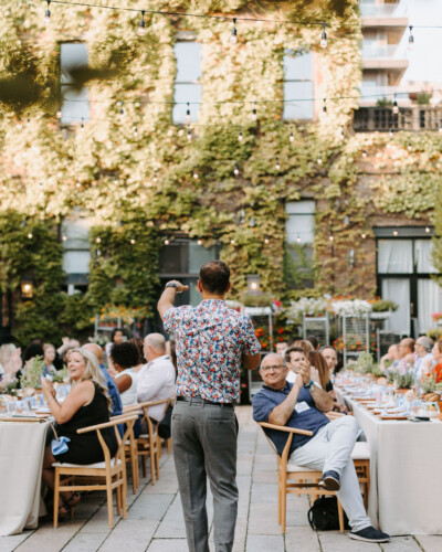 A person stands speaking to a group of people seated at tables in an outdoor garden setting.