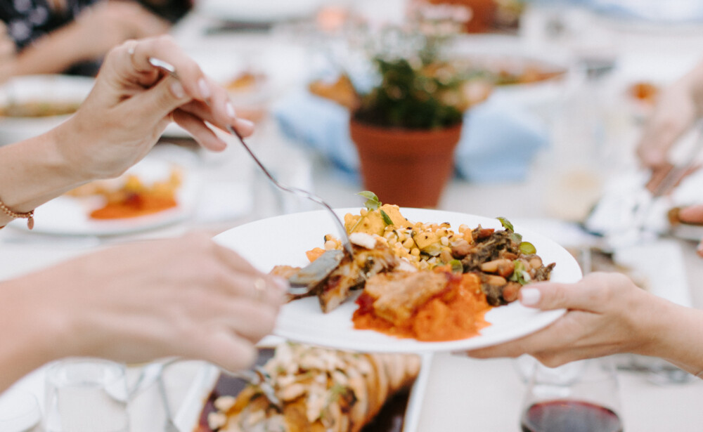 People share and serve food at a long outdoor dining table set with plates, drinks, and potted plants.