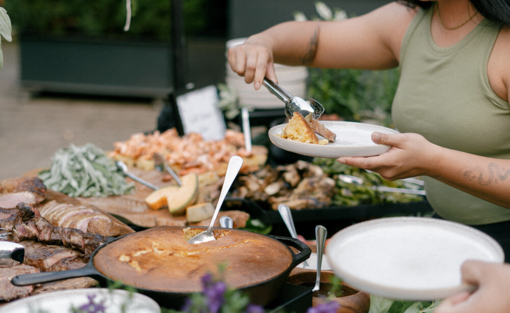A woman serves food from a buffet table outdoors, surrounded by flowers and various dishes.