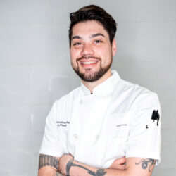 Smiling chef with tattoos, wearing a white chef coat, stands with arms crossed against a plain background.