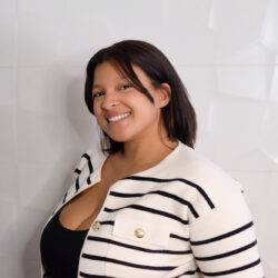 A woman smiling, wearing a striped jacket and black top, standing against a white textured wall.