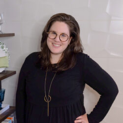 Woman with glasses and long brown hair smiles, wearing a black dress and long necklace, standing indoors.