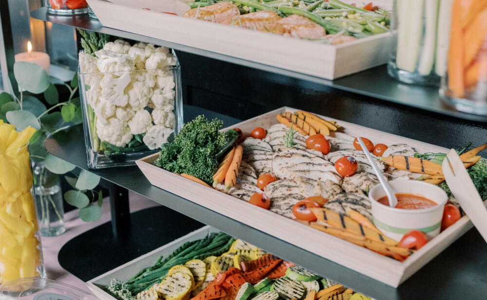 A buffet table with trays of grilled vegetables, fresh veggie sticks, dip, and salad bowls.
