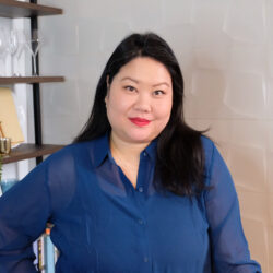 Woman with long dark hair in a blue shirt smiling, standing indoors near shelves with glasses and books.