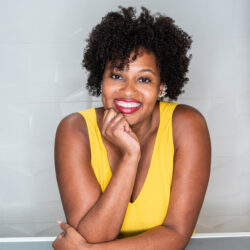A woman in a yellow sleeveless top smiles with her hand resting on her chin against a light background.