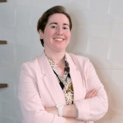 A person in a light pink blazer stands smiling with arms crossed in front of a white textured wall.
