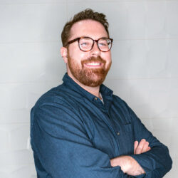 A smiling man with glasses and a beard stands with arms crossed, wearing a blue shirt against a light background.