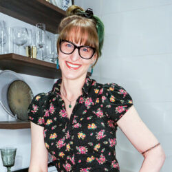 Woman with glasses wearing a floral shirt, smiling in a modern kitchen with shelves and glassware behind her.