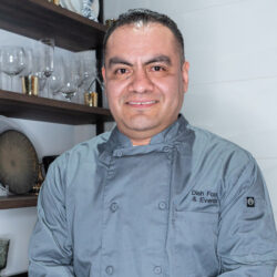 A man in a gray chef coat stands smiling in front of shelves with glassware and decorative items.