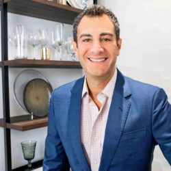 Smiling man in a blue suit stands in front of shelves with glassware and decorative plates.