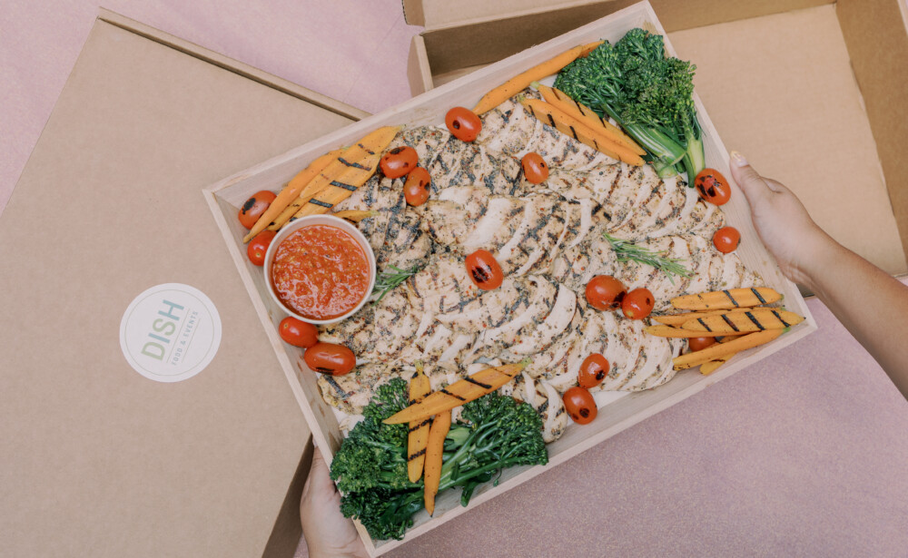 A person holds a box of grilled chicken, carrots, broccoli, cherry tomatoes, and a cup of red sauce.