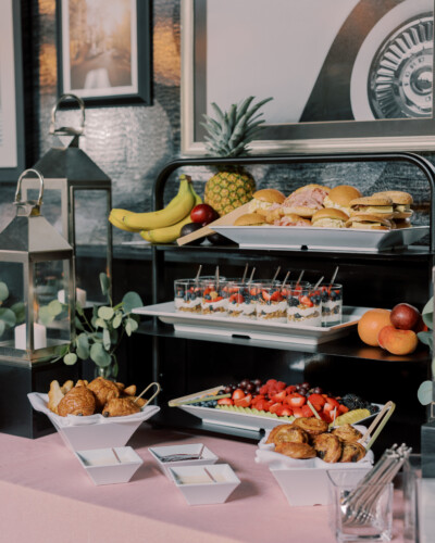 Buffet table with sandwiches, fruit, parfaits, pastries, and decorative lanterns on a pink tablecloth.