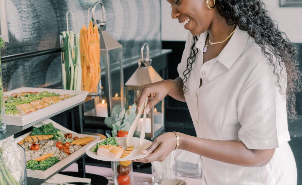 A woman in a white dress serves food from a buffet table with salads and vegetables.