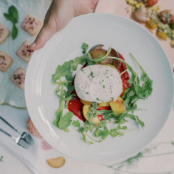 Hand holding a plate of burrata cheese with arugula, tomatoes, and greens; other dishes on table.