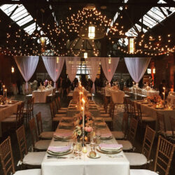 Elegant wedding reception hall with string lights, long tables, floral centerpieces, and draped white curtains.