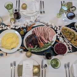 A holiday dinner table set with sliced ham, mashed potatoes, stuffing, cranberry sauce, and festive decorations.