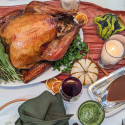 Roast turkey on a platter with herbs, surrounded by candles, drinks, gourds, and a bowl of gravy.