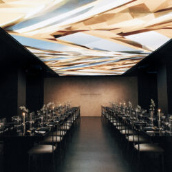 Elegant dining room with two long tables, black chairs, and a geometric, illuminated ceiling design.