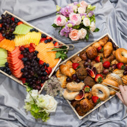 Two trays with assorted fruit and pastries on gray fabric with flowers; a hand reaches for a bagel.