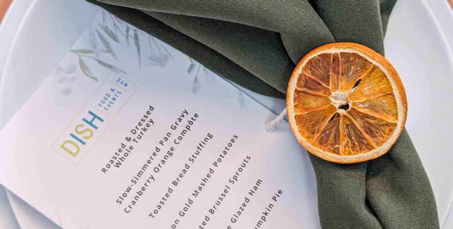 Place setting with a green napkin, dried orange slice, and printed menu on a white plate with a brown rim.