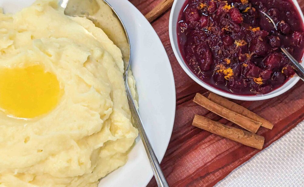 Bowl of mashed potatoes with butter, cranberry sauce, small pumpkins, and cinnamon sticks on a festive table.