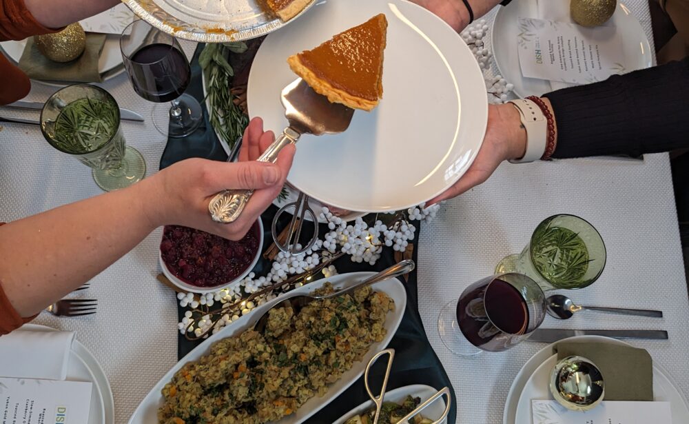 Hands serve pumpkin pie at a festive table with Brussels sprouts, stuffing, drinks, and candles.