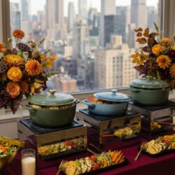 Buffet with flowers and food trays in front of a window showing a city skyline with tall buildings.